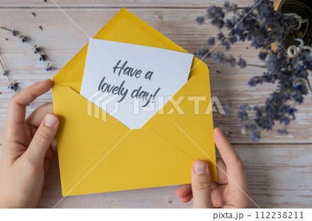 Female hands taking paper card note with text HAVE A LOVELY DAY from yellow envelope. Lavender flower. Top view, flat lay. Concept of mental spiritual health Female hands taking paper card note with text HAVE A LOVELY DAY from yellow envelope. Lavender flower. Top view, flat lay. Concept of mental spiritual health 112238211