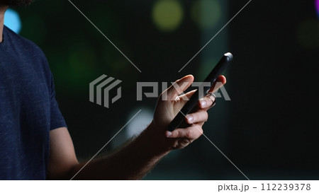 Modern person on smartphone during stroll late at night, looking around city centre with illuminated skyscrapers. Casual man texting on phone and walking downtown. Handheld shot. Close up. 112239378