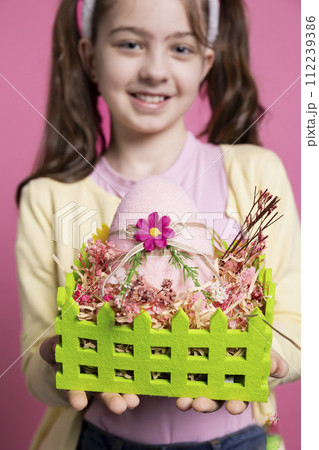 Positive excited small kid showing her handmade easter eggs in a colored basket, feeling proud of her arrangement. Young child with bunny ears holding festive decorations, april celebrations. 112239386