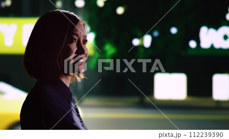 Asian adult admiring skyscrapers on walk and looking around downtown district, enjoying evening stroll around city. Woman smiling and relaxing on urban night time promenade in town. Handheld shot. 112239390