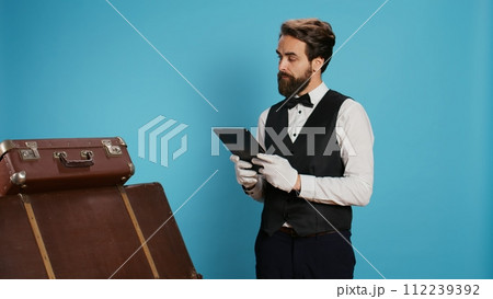 Classy doorman porter reviewing tablet, in charge of baggage for safekeeping. Bellboy presenting his commitment in providing outstanding concierge service, symbolizing his occupation at chic hotel. 112239392