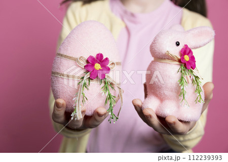 Cute small kid presenting her handmade decorated ornaments, adorable pink egg and a stuffed rabbit in studio. Little girl smiling and feeling excited about easter celebration event. Close up. 112239393
