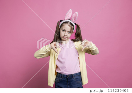 Negative displeased young kid showing thumbs down sign in studio, acting disappointed and sad before easter celebration. Mad angry small child being confident with her dislike symbol on camera. 112239402
