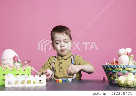 Delighted young kid coloring eggs and ornaments with watercolors as he prepares artistic creations for Easter Sunday festivities. Playful toddler having fun in the studio with tie dye painting. Delighted young kid coloring eggs and ornaments with watercolors as he prepares artistic creations for Easter Sunday festivities. Playful toddler having fun in the studio with tie dye painting. 112239470