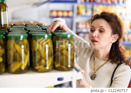 Adult female shopper in casual clothes chooses pickled cucumbers in grocery store 112240757