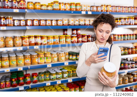 Female shopper scans barcode on jar of sauerkraut using a smartphone while shopping in grocery store 112240902