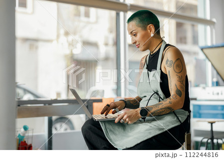 Positive female designer sitting with laptop on background of print machine in workshop 112244816