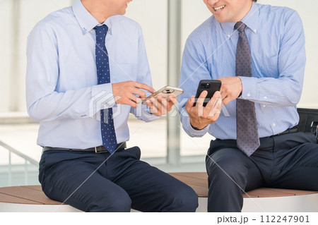 オフィスの休憩所・ベンチでスマホを見ながら笑顔で会話する男性ビジネスマン（上司・部下） 112247901