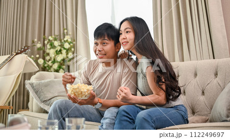 A cheerful Asian couple is watching an exciting sports match or game show together at home. A cheerful Asian couple is watching an exciting sports match or game show together at home. 112249697