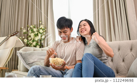 A cheerful Asian couple is watching an exciting sports match or game show together at home. 112249698