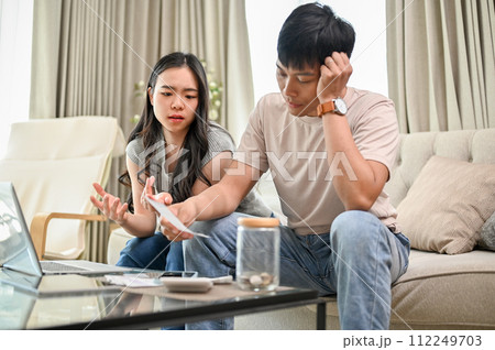 An unhappy young Asian couple is arguing about high domestic bills on a sofa in the living room. An unhappy young Asian couple is arguing about high domestic bills on a sofa in the living room. 112249703