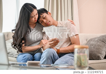 A stressed young married couple is sitting on a sofa, concerned about their invoices. 112249721