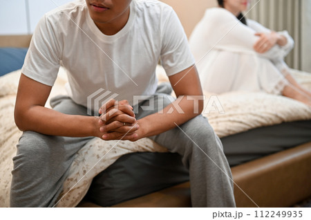 An angry, unhappy Asian husband sitting on bed separately with his wife after their argument. An angry, unhappy Asian husband sitting on bed separately with his wife after their argument. 112249935