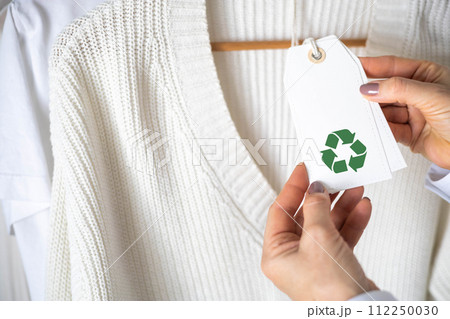 Female hands hold a label from white knitted clothing, which depicts the Recycling sign. 112250030