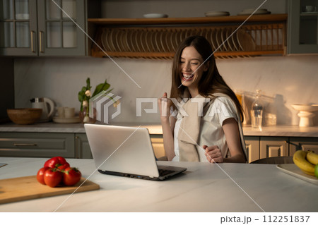 Joyful Young Woman Celebrating Success at Home With Laptop in Kitchen Joyful Young Woman Celebrating Success at Home With Laptop in Kitchen 112251837