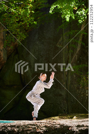 Woman practicing yoga outdoors in forest. Barefoot female on yoga mat surrounded by trees and large rocks, which suggests peaceful, natural environment ideal for meditation or yoga practice. Woman practicing yoga outdoors in forest. Barefoot female on yoga mat surrounded by trees and large rocks, which suggests peaceful, natural environment ideal for meditation or yoga practice. 112252954