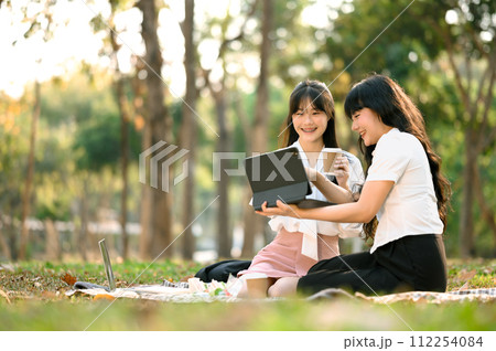 Smiling Asian female colleagues using digital tablet and relaxing in the park 112254084