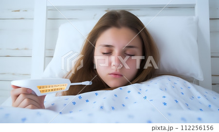 A teenage girl is lying in bed, holding a thermometer in her ha A teenage girl is lying in bed, holding a thermometer in her ha 112256156