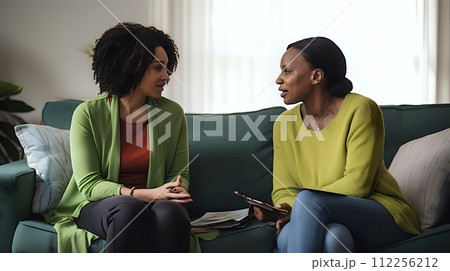 Two women are sitting on a couch having a conversation. One woma 112256212