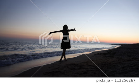 Woman standing with open arms on seashore at sunset 112256267