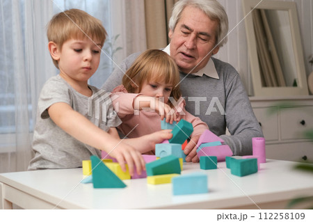 Grandfather playing toys with grandson and young granddaughter indoor. Old man grandpa studying with his grandchildren at weekend. Senior male having fun with kids. Happy multigeneration family video 112258109