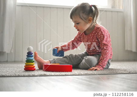 Kid girl play toys at home, kindergarten or nursery. Baby child playing playthings cubes and wood puzzle, pyramid sitting on the floor. Happy preschool education indoor. 3 years old little toddler. 112258116