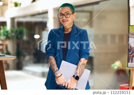 A woman with green hair and tattoos smiles while holding a laptop in formal wear 112259151