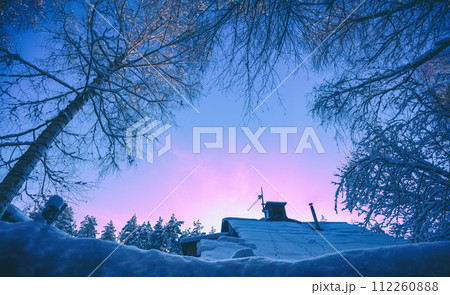 Winter landscape in the evening. View through the trees to a snowy roof with a chimney. Village in the forest in snowy winter at sunset 112260888