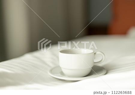Coffee or tea cup on the bed. Hotel room or bedroom Interior, morning time. Concept of easy breakfast. One white ceramic cup. 112263688