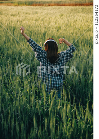 Young pretty woman in red summer dress and straw hat walking on yellow farm field with ripe golden wheat enjoying warm evening. 112264172