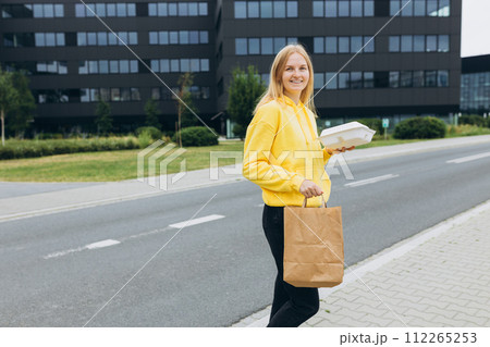 Happy woman receiving fresh takeaway lunches, standing near the office building. Delivery in any weather around the clock to the client. Concept of rest and break on job. Happy woman receiving fresh takeaway lunches, standing near the office building. Delivery in any weather around the clock to the client. Concept of rest and break on job. 112265253
