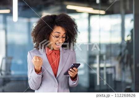 Close-up photo of a happy young African-American woman in a suit standing in the office, looking at the phone screen and happy to receive news, showing a thumbs-up gesture with her hand. 112268626
