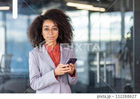 Close-up portrait of a serious young African-American businesswoman standing in the office in a suit and glasses, looking at the camera and holding a mobile phone. Close-up portrait of a serious young African-American businesswoman standing in the office in a suit and glasses, looking at the camera and holding a mobile phone. 112268627