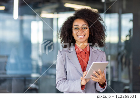 Portrait of a smiling and successful young African American woman in a suit standing in a modern office, holding a tablet and looking at the camera. 112268631