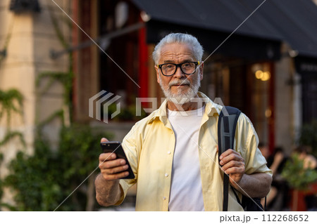 Portrait of a gray-haired senior man dressed in casual clothes and glasses, holding a backpack and a mobile phone, standing on a city street and looking at the camera with a smile. 112268652