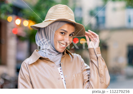 Portrait of attractive muslim woman wearing hijab and touching straw hat with playful facial expression. Delighted lady combining headwear and feeling ready for vacation season in stylish outfit. 112270256
