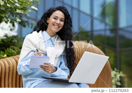 Successful diverse entrepreneur sitting on wooden bench and making notes in daybook while holding computer on laps. Smiling female writing to do list by hand for not skipping important tasks. Successful diverse entrepreneur sitting on wooden bench and making notes in daybook while holding computer on laps. Smiling female writing to do list by hand for not skipping important tasks. 112270263