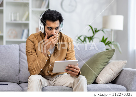 Shocked muslim guy in white headphones covering mouth with hand while looking at digital tablet screen in living room. Amazed man watching thrilling horror movie and getting impressed by epic moment. Shocked muslim guy in white headphones covering mouth with hand while looking at digital tablet screen in living room. Amazed man watching thrilling horror movie and getting impressed by epic moment. 112270323