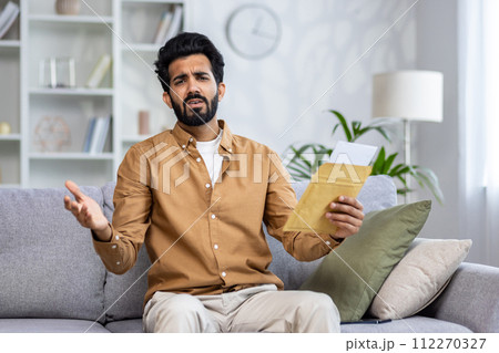 Confused hispanic male spreading hands with open envelope while looking at camera with disappointed facial expression. Worried man in shirt receiving bad news and unpleasant notification from bank. 112270327