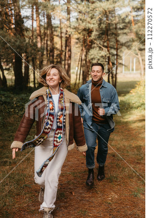 Happy couple is having fun in the middle of the forest, a smiling woman is running with her husband in an autumn park. Happy couple is having fun in the middle of the forest, a smiling woman is running with her husband in an autumn park. 112270572