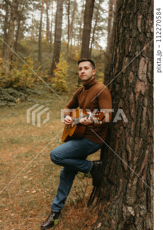 Caucasian adult man plays guitar and leaning on tree in the autumn park Caucasian adult man plays guitar and leaning on tree in the autumn park 112270584
