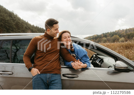 Happy man leaning to a car and holding hands of cheerful woman that sitting inside, adult couple enjoying road trip in autumn 112270586
