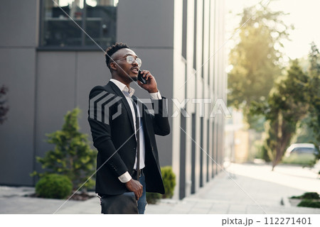Serious young businessman in black suit standing and talking on cell phone near business center Serious young businessman in black suit standing and talking on cell phone near business center 112271401