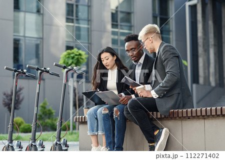 Confident international co workers sitting on wooden bench in front of office and discuss results of joint work. Confident international co workers sitting on wooden bench in front of office and discuss results of joint work. 112271402
