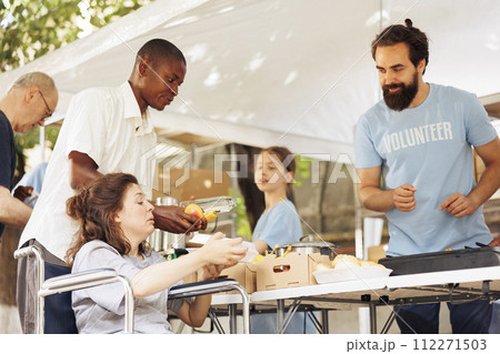 Poor caucasian female wheelchair user receives free food and nourishments at outdoor food bank. Male charity worker in blue t-shirt provides fresh fruits and warm meals to needy and less privileged. Poor caucasian female wheelchair user receives free food and nourishments at outdoor food bank. Male charity worker in blue t-shirt provides fresh fruits and warm meals to needy and less privileged. 112271503