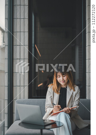 Engrossed in her thoughts, a woman pensively jots down notes beside her laptop in a modern workspace, a moment of deep concentration captured amidst her creative process. 112272120