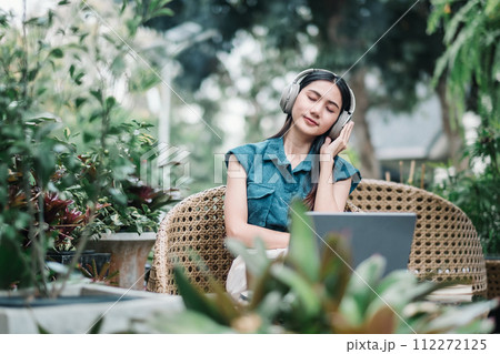 A serene young woman wearing headphones, listening to music while sitting with a laptop in a lush garden setting. A serene young woman wearing headphones, listening to music while sitting with a laptop in a lush garden setting. 112272125