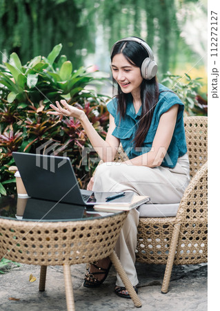 A cheerful young woman with headphones sits at a rattan chair, engaging in a video call on her laptop amidst lush greenery. A cheerful young woman with headphones sits at a rattan chair, engaging in a video call on her laptop amidst lush greenery. 112272127