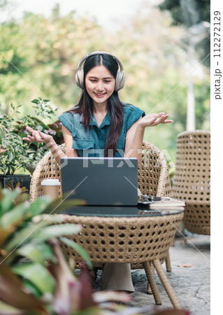 A cheerful young woman with headphones sits at a rattan chair, engaging in a video call on her laptop amidst lush greenery. 112272129