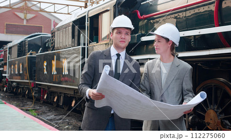 Young caucasian engineer man and woman in suit checking train looking blueprint in station. Young caucasian engineer man and woman in suit checking train looking blueprint in station. 112275060